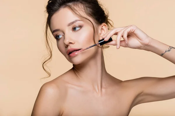 Young woman with naked shoulders holding brush of lip gloss isolated on beige