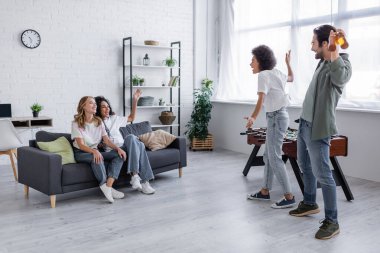 Mutlu lezbiyen çift kanepede oturuyor ve bira şişeleriyle ırklar arası arkadaşlara bakıyor.