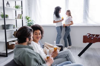 Çok ırklı lezbiyen çift ellerinde bira şişeleriyle ön planda bulanık arkadaşlarının yanında dikiliyorlar. 