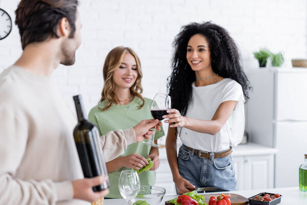 blurred man giving glass of wine to cheerful african american woman near blonde friend in kitchen