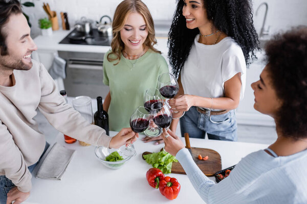 high angle view of happy multiethnic friends clinking glasses of red wine in kitchen 