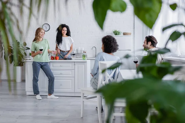 前景に植物がぼやけている現代のキッチンで話している幸せな多民族の友人 — ストック写真