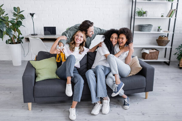 happy man holding bottles of beer and hugging interracial women on sofa