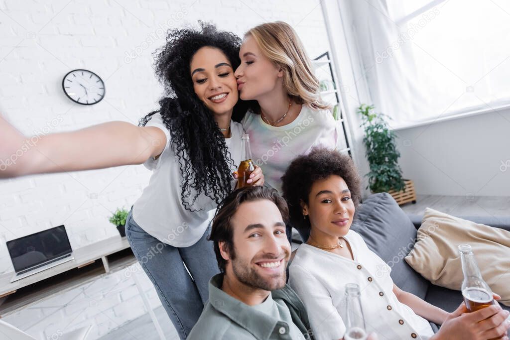 happy blonde woman kissing cheek of african american girlfriend near cheerful friends in living room