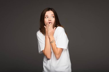 scared and shocked woman covering mouth with hands while looking away isolated on dark grey clipart