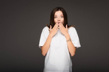shocked woman covering mouth with hands while looking at camera isolated on dark grey clipart