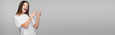 astonished woman pointing with fingers while looking at camera isolated on grey, banner clipart