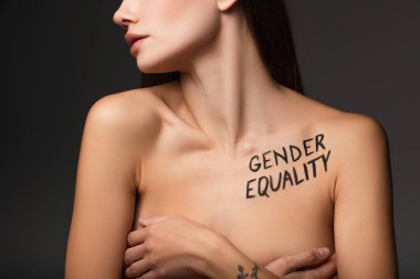 partial view of young woman with gender equality inscription on body covering breast with hands isolated on dark grey clipart