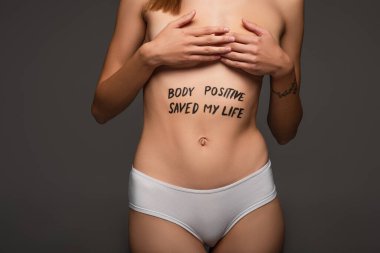 partial view of woman with body positive saved my life lettering on belly covering breast with hands isolated on dark grey clipart