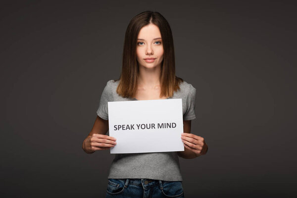 young woman holding card with speak your mind phrase isolated on grey
