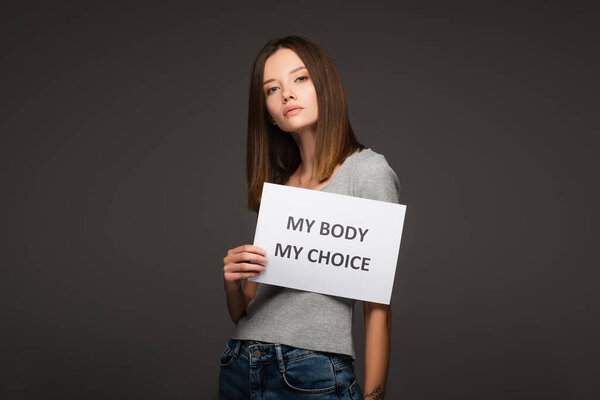 brunette woman looking at camera while holding card with my body my choice inscription isolated on grey