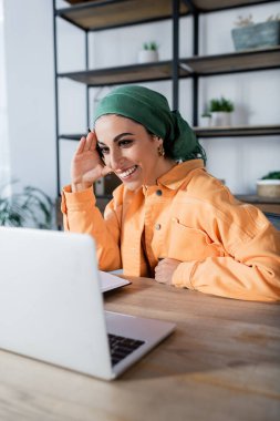 Evdeki online seminerde dizüstü bilgisayarın yanında gülümseyen neşeli Müslüman kadın.