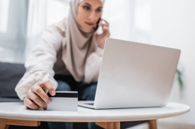Bilgisayarın yanında cep telefonuyla konuşan Müslüman kadının elindeki kredi kartının seçici odak noktası.
