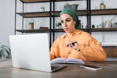 muslim woman in headkerchief and orange jacket sitting with pen near blurred notebook clipart