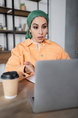 surprised muslim woman in headkerchief pointing at blurred laptop near coffee to go clipart