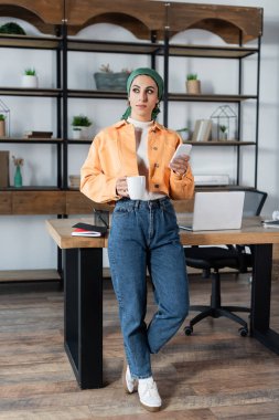 full length view of muslim woman with tea cup and smartphone standing near desk at home clipart