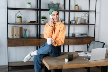 happy muslim student with tea cup talking on smartphone while sitting on desk near laptop clipart