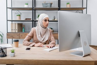 Akıllı telefonun yanında bilgisayar başında çalışan genç Müslüman kadın. Boş ekranı ve kahvesi var.