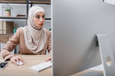 Bulanık bilgisayar monitörü ve evde boş ekranlı akıllı telefon ile çalışan Müslüman kadın.