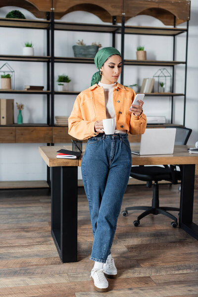 Вид женщины в полный рост, использующей смартфон, стоя с чашкой чая в гостиной