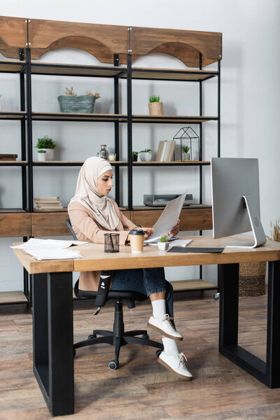muslim woman looking at document while sitting near computer at home