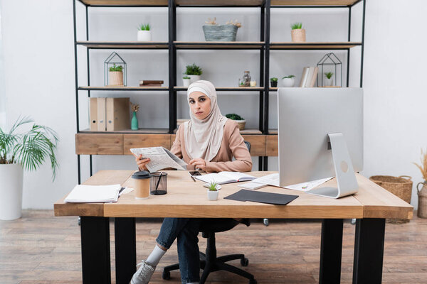muslim woman holding newspaper and looking at camera near computer on work desk at home