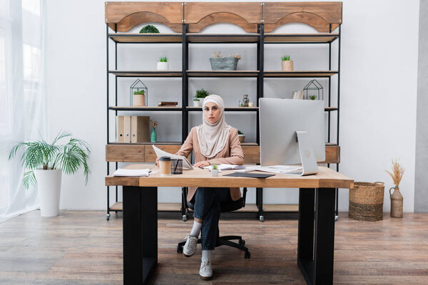muslim woman looking at camera while sitting near computer and documents in home office
