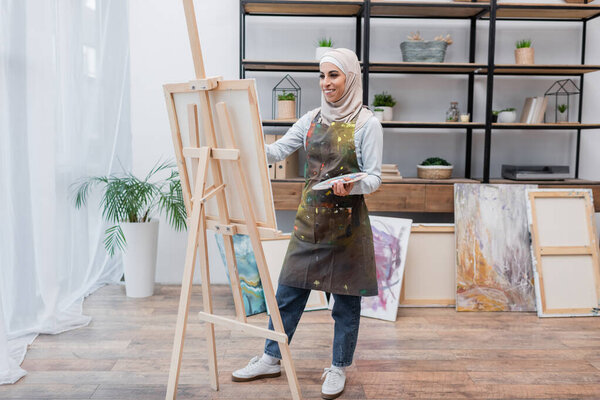 full length view of happy muslim woman in apron drawing on easel at home