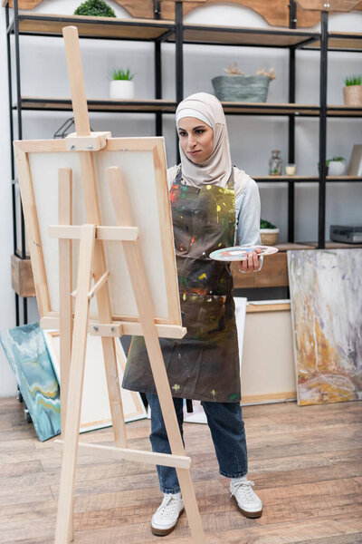 full length view of muslim woman painting on easel in home studio