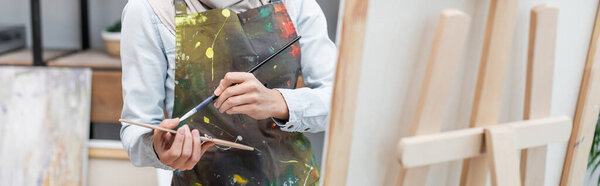 partial view of artist with palette and paintbrush near easel, banner