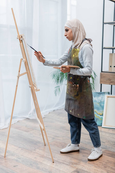full length view of arabian woman in hijab and apron drawing at home