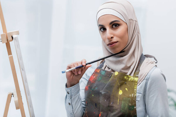 young muslim woman holding paintbrush while looking at camera near easel