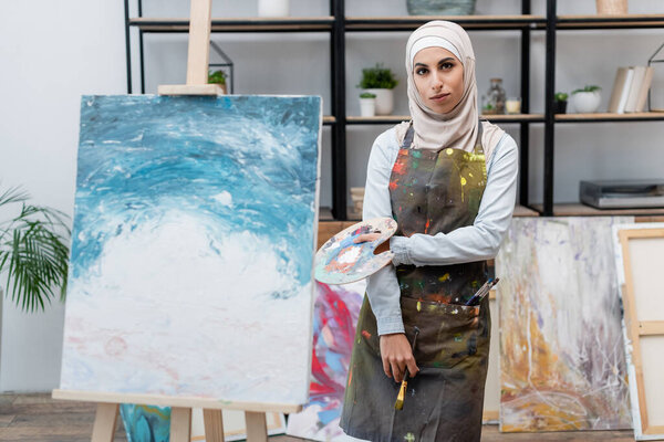 young arabian woman with palette and paintbrush looking at camera near picture on easel