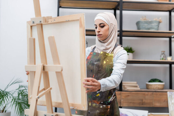 young muslim woman in hijab and apron near easel in home studio