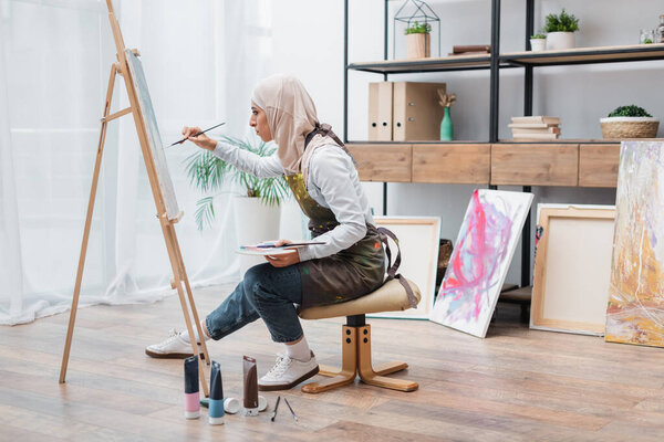 side view of muslim woman sitting on chair and drawing on easel at home