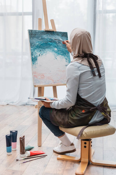 back view of muslim woman drawing on canvas near paintbrushes and paint tubes on floor