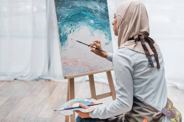 young muslim woman in hijab painting on easel at home