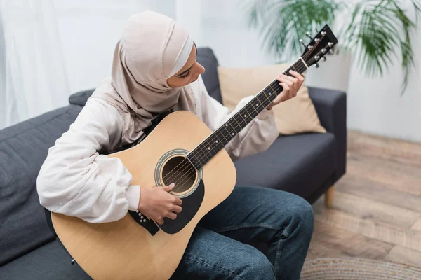 Ung Muslimsk Kvinde Sidder Sofaen Derhjemme Spiller Guitar - Stock-foto