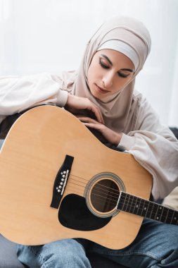 Akustik gitara yaslanmış, evde oturan tesettürlü genç Arap kadın.