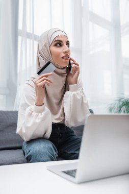 Bilgisayarın yanında cep telefonuyla konuşan Arap bir kadın kredi kartı tutuyor.