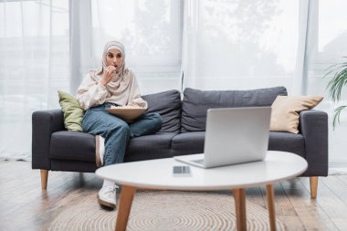 Müslüman kadın kanepede oturuyor ve dizüstü bilgisayardan film izlerken patlamış mısır yiyor.
