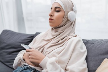 Kulaklıklı müslüman kadın akıllı telefon tutarken kapalı gözlerle müzik dinliyor.