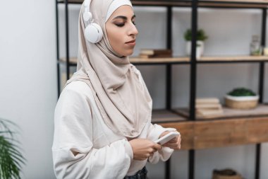 Müslüman kadın, oturma odasında akıllı telefonuyla dikilmiş kulaklıkla müzik dinliyor.