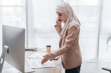 Bilgisayarın yanında cep telefonuyla konuşan müslüman kadının grafiklere baktığı yan görünüm