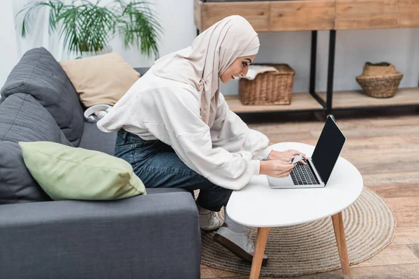 坐在沙发上用空白屏幕在笔记本电脑上打信用卡的穆斯林妇女的侧视图 — 图库照片
