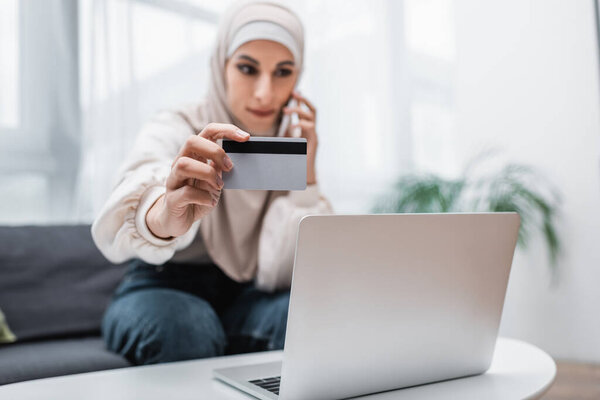 selective focus of credit card in hand of blurred arabian woman calling on smartphone near laptop