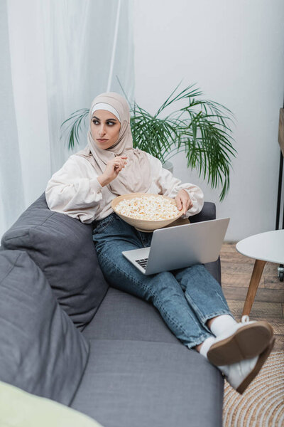 muslim woman looking away while sitting on couch with popcorn and laptop