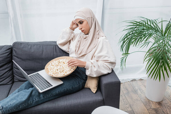 pensive arabian woman with bowl of popcorn watching movie on laptop near green potted palm