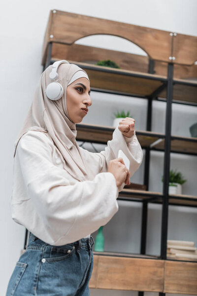 young muslim woman in headphones holding smartphone and dancing at home