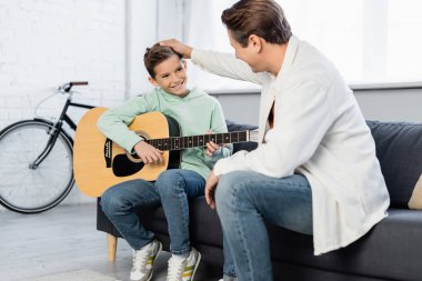 Gülümseyen baba koltukta akustik gitarla oğlunun saçına dokunuyor. 